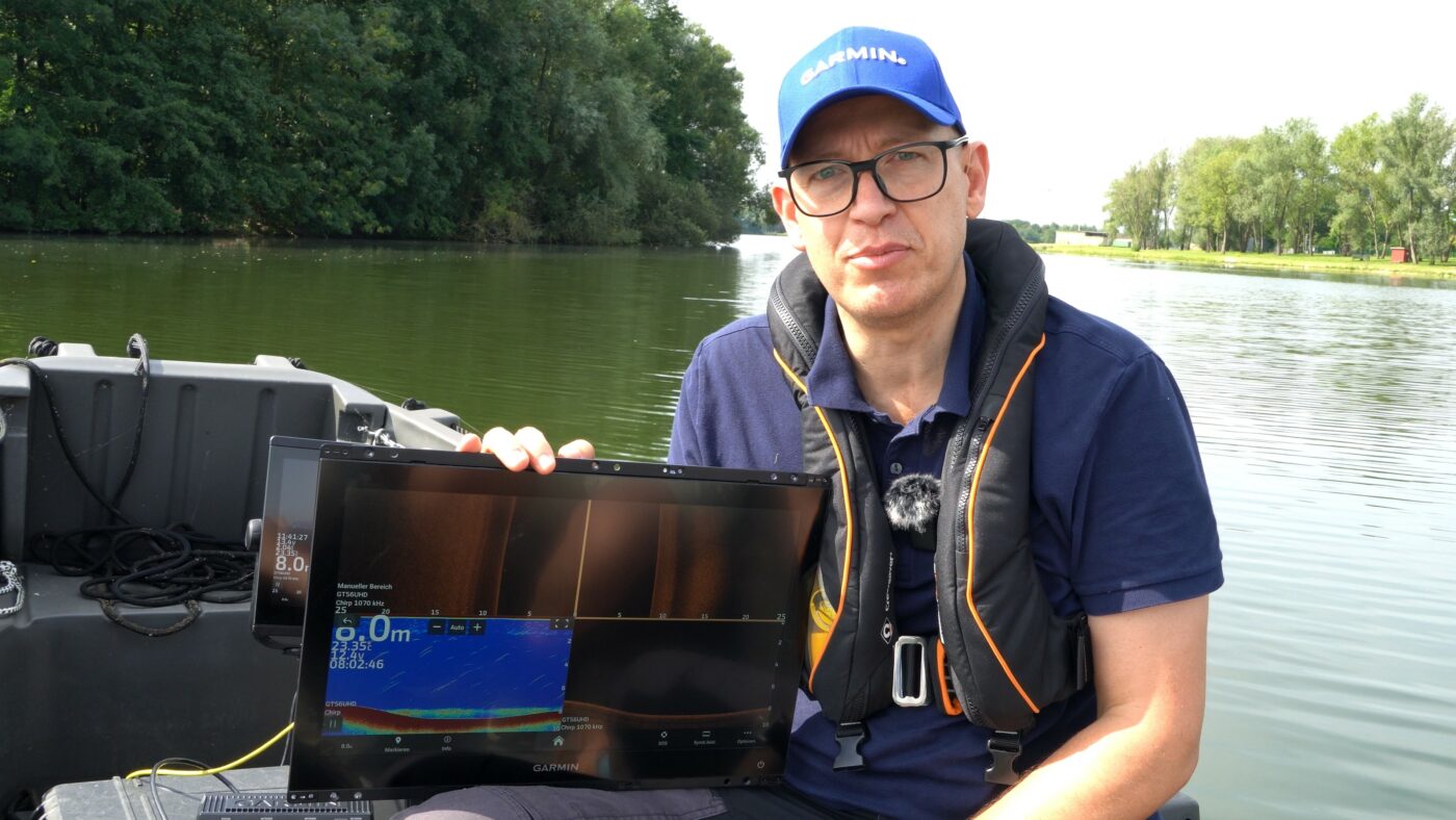 Eine Person auf einem Boot hält ein Echolot-Gerät. Im Hintergrund ist ein Fluss mit Bäumen zu sehen.