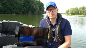 Eine Person auf einem Boot hält ein Echolot-Gerät. Im Hintergrund ist ein Fluss mit Bäumen zu sehen.